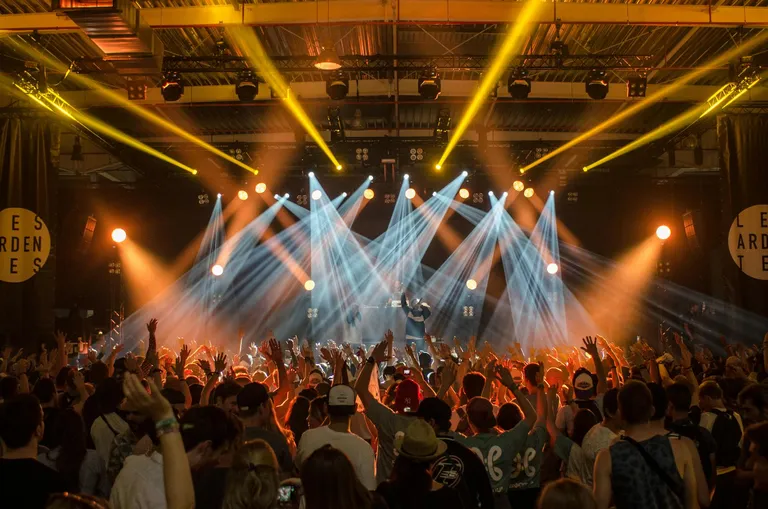 A lively concert scene with a crowd raising their hands towards the stage, illuminated by vibrant lighting effects.