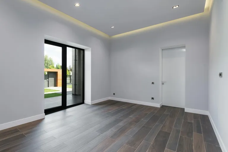 A modern empty room with large glass doors, wooden flooring, and minimalistic design, showcasing architectural lighting.