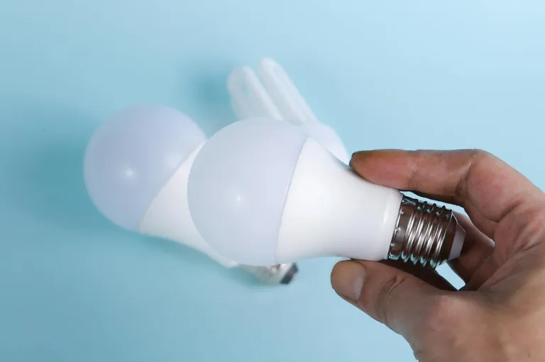 A person holding a modern LED light bulb with a white bulb shape, alongside another bulb in the background, on a light blue surface.