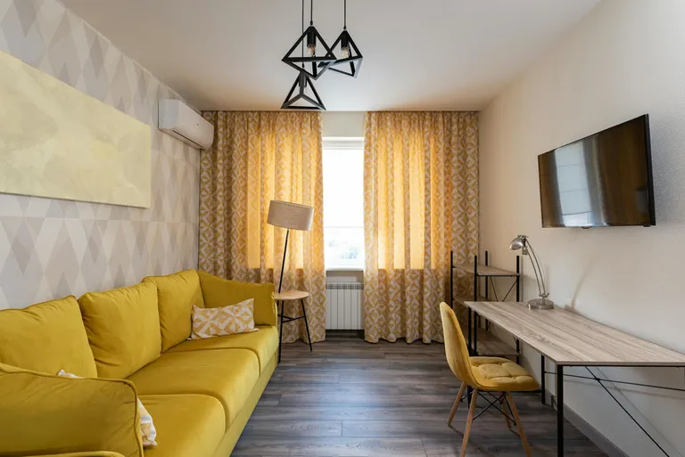 A modern and stylish small living room with a bright yellow couch, wooden flooring, and elegant lighting fixtures, featuring patterned curtains and a sleek desk setup.