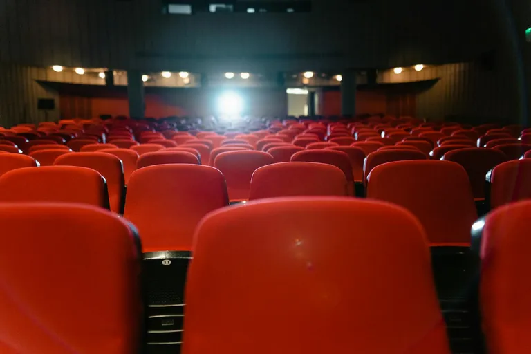 A view of red theater seats leading towards the stage of a darkened auditorium, with a focused light shining from the back, creating an inviting atmosphere for an event.